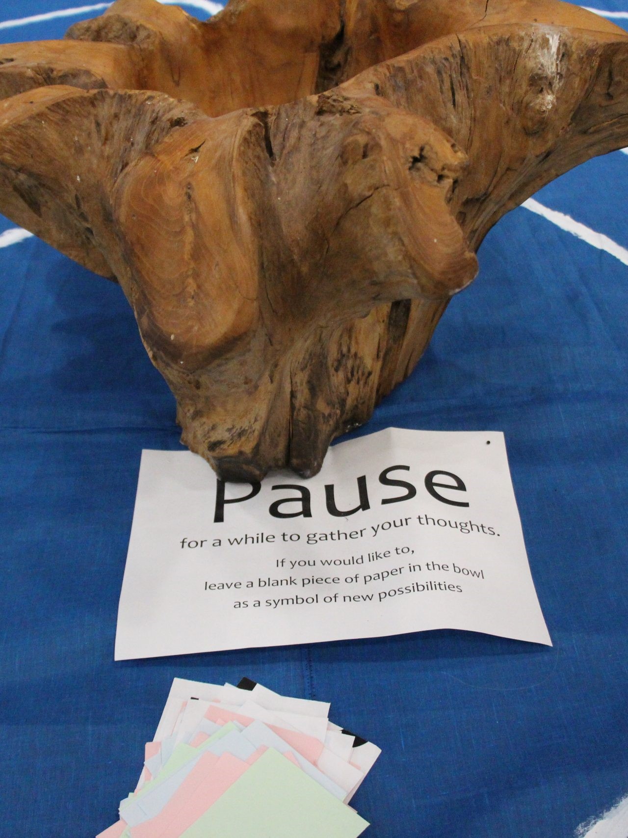 A wooden bowl. A sign in front of it reads: "Pause for a while to gather your thoughts. If you would like to, leave a blank piece of paper in the bowel as a symbol of new possibilities.”