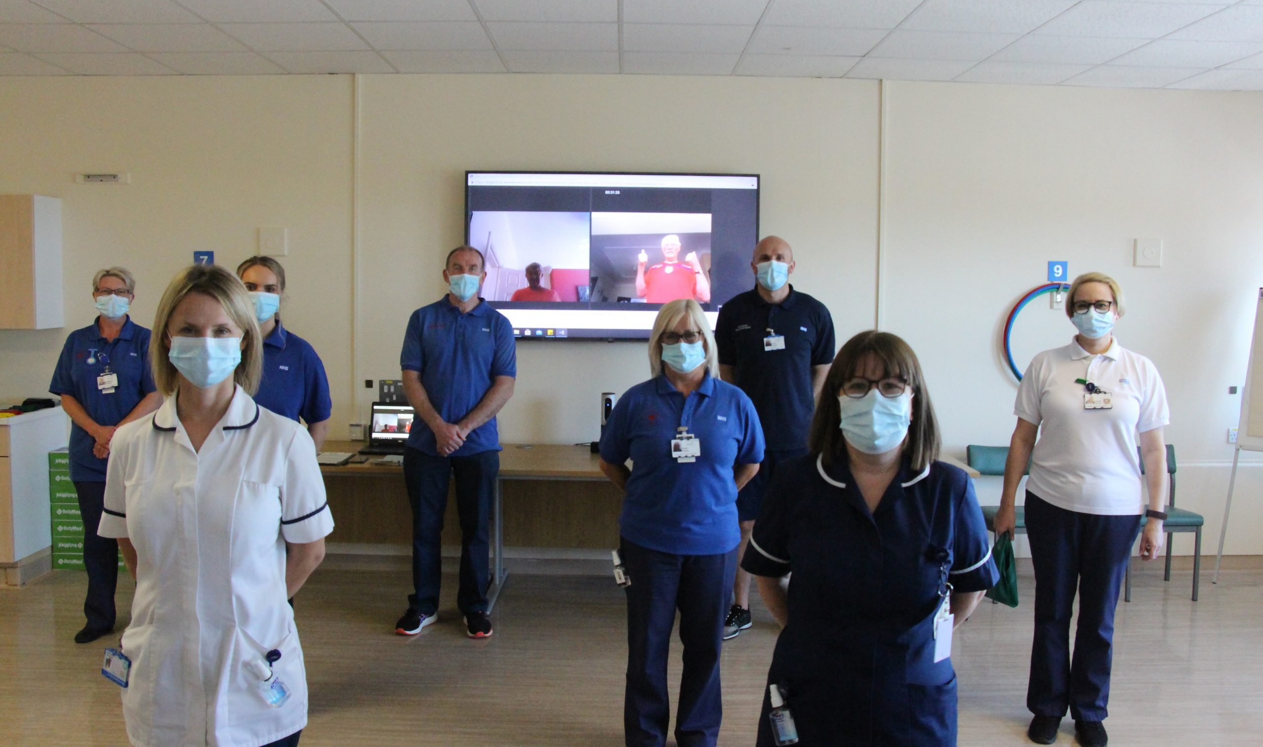 The team stand in front of a TV screen. The screen shows two patients giving thumbs up.