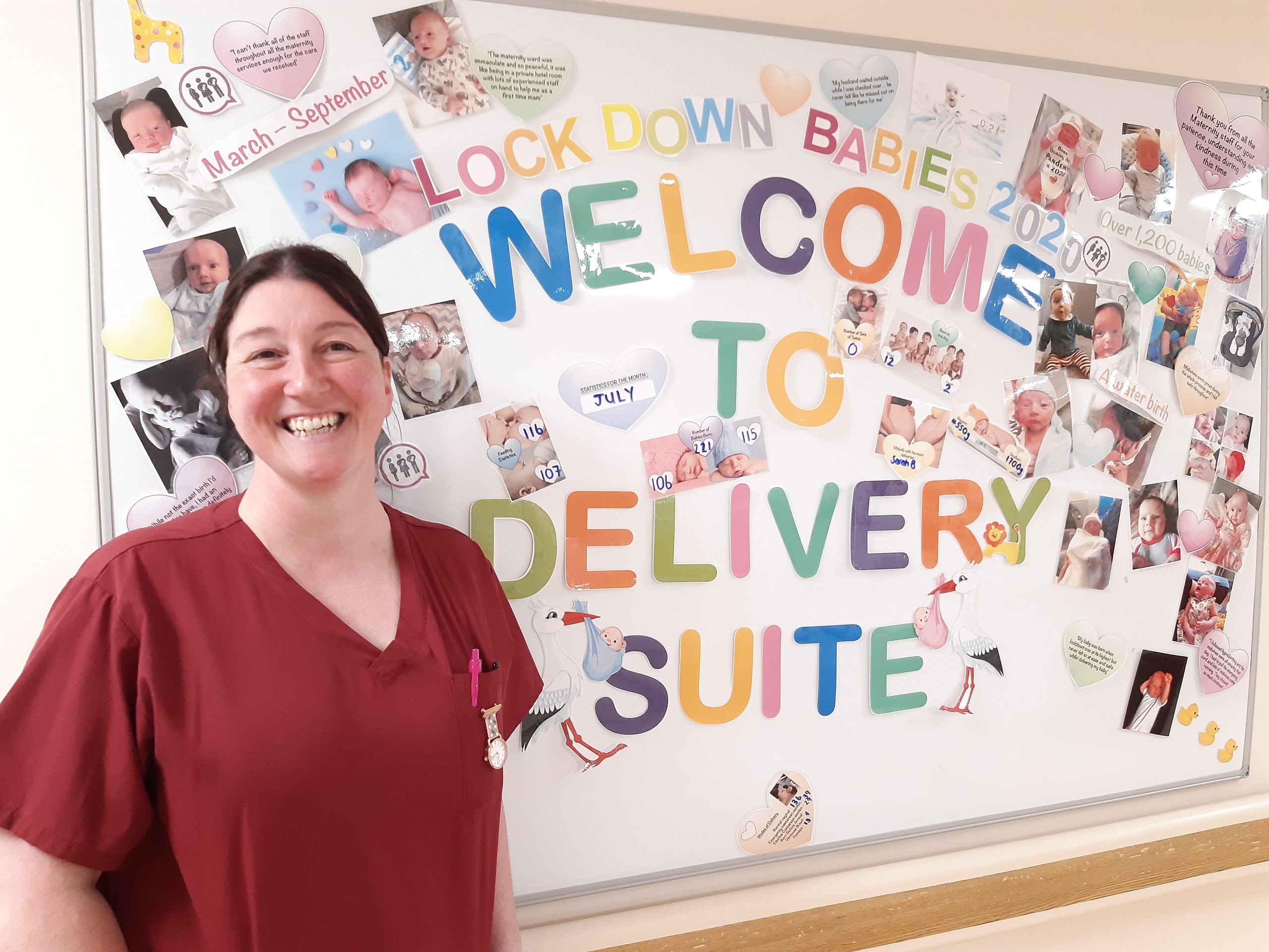 Nicola stands in front of a white board. Letters spell out "Lockdown babies 2020: Welcome to delivery suite". There are photos of babies on the board.