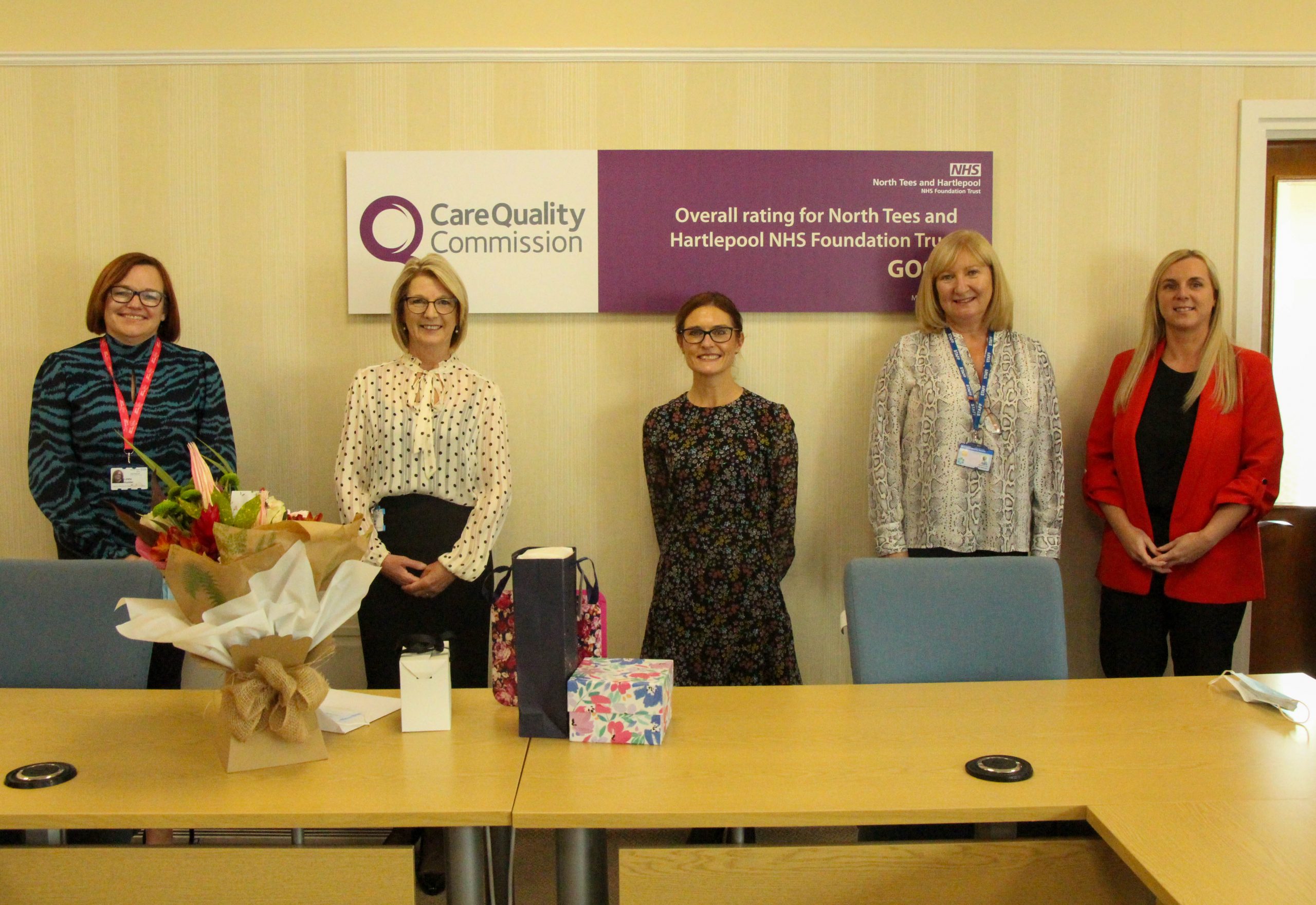 Lynne stands with colleagues. On the table in front of her are flowers and gifts.
