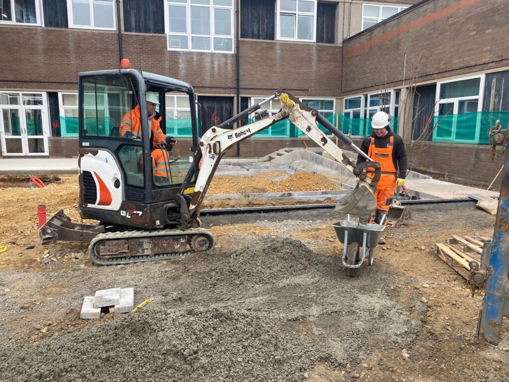 Two construction workers on the building site. One sits in a crane and the other holds a wheelbarrow.