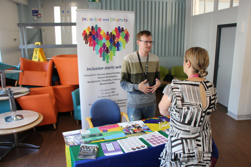 A lady talks to a man hosting a stall. There are information leaflets on the table. A banner behind the man reads "Inclusion and diversity".