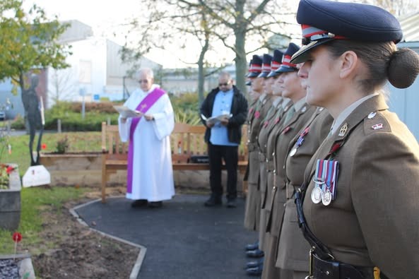 A garden with poppies. Members of the Armed Forces stand in uniform. A chaplain reads from a book.