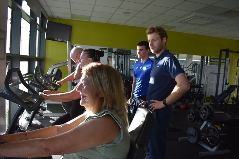 Two people on exercise bikes. Hospital staff and students stand behind them.