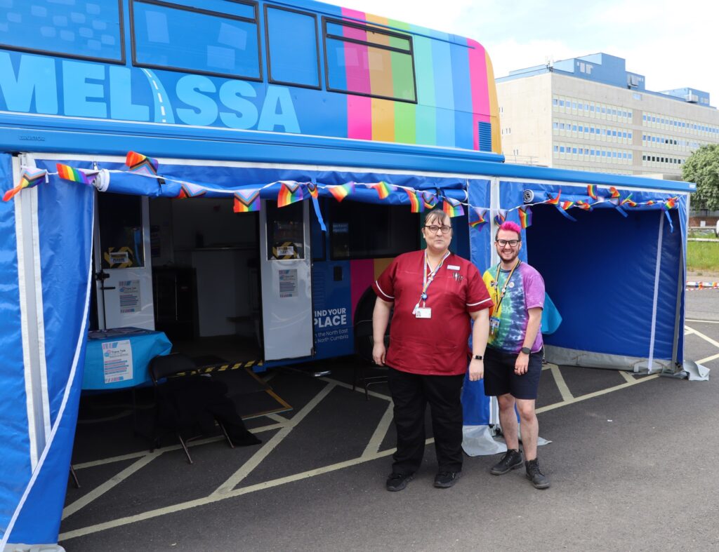 Two members of staff stand in front of a blue bus.