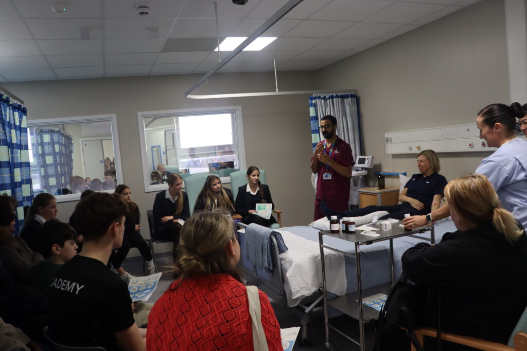 A person lies in a hospital bed. A doctor stands to them and talks to a group of children and teenagers.