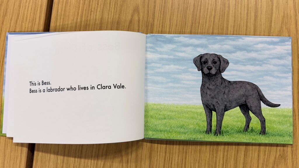 Pages in the book. One page reads: "This is Bess. Bess is a labrador who lives in Clara Vale." The opposite page is an illustration of a black labrador standing on grass.