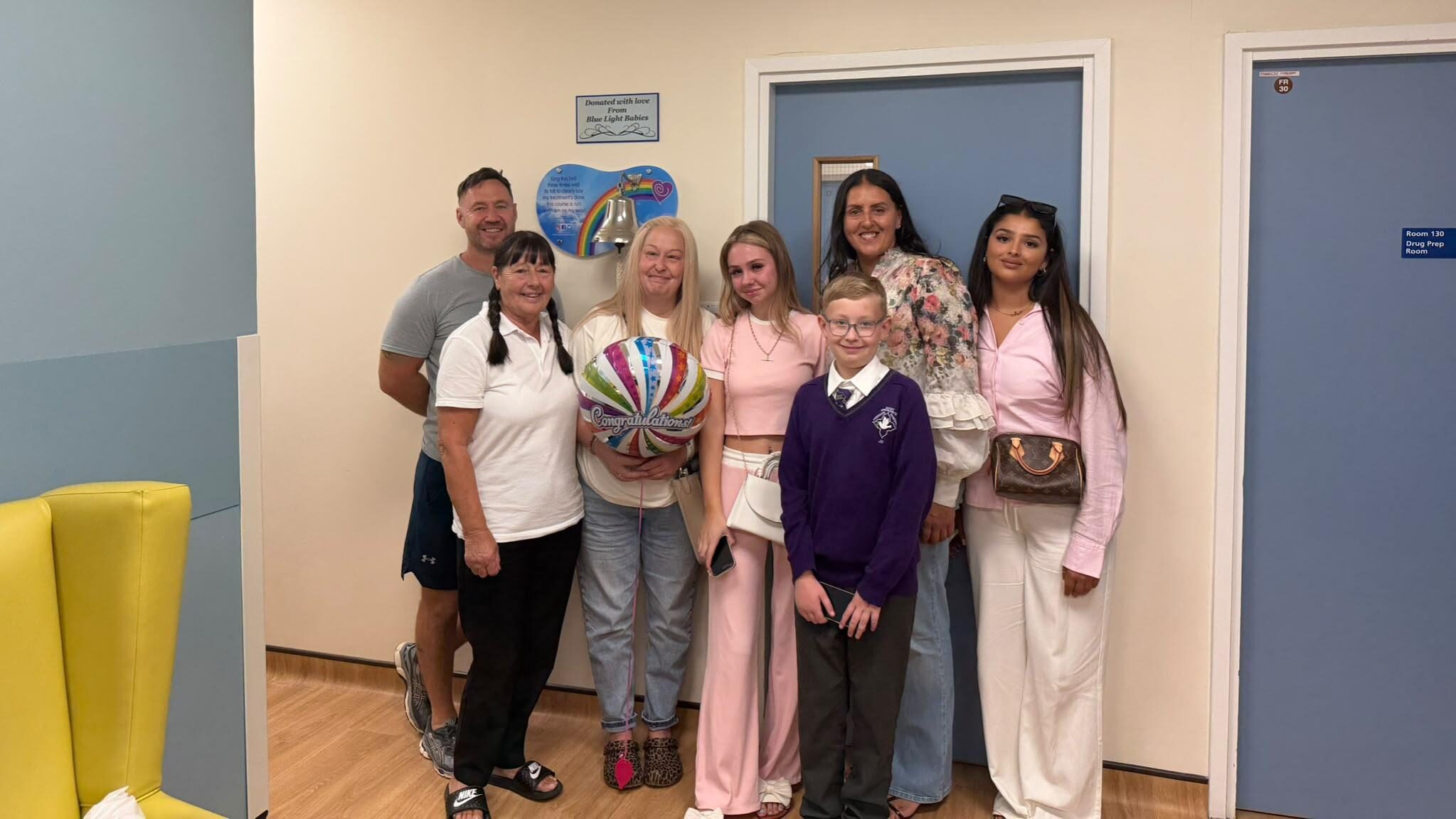 Lisa and her family in the chemotherapy unit. They stand in front of a bell. Lisa holds a balloon which reads 'Congratulations'.