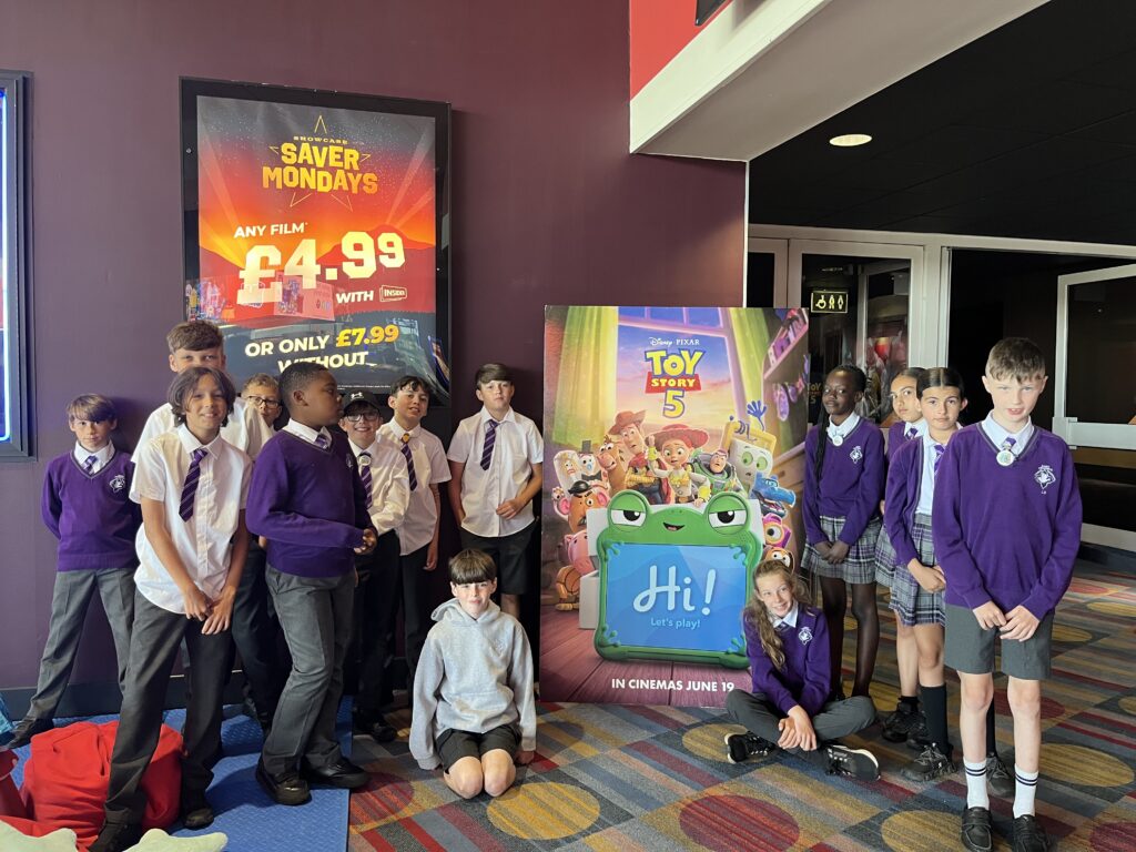 A group of children in school uniform in a cinema foyer. They stand with a poster for Toy Story 5.