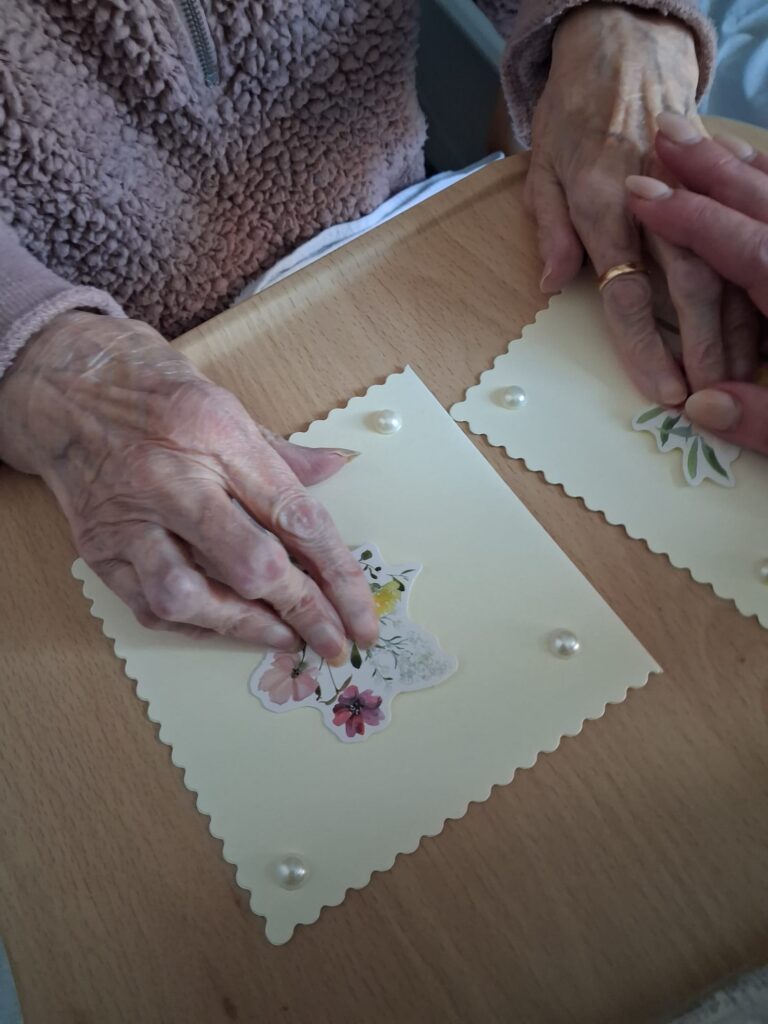 A pair of elderly hands, resting on two homemade cards. Someone holds one of the hands.