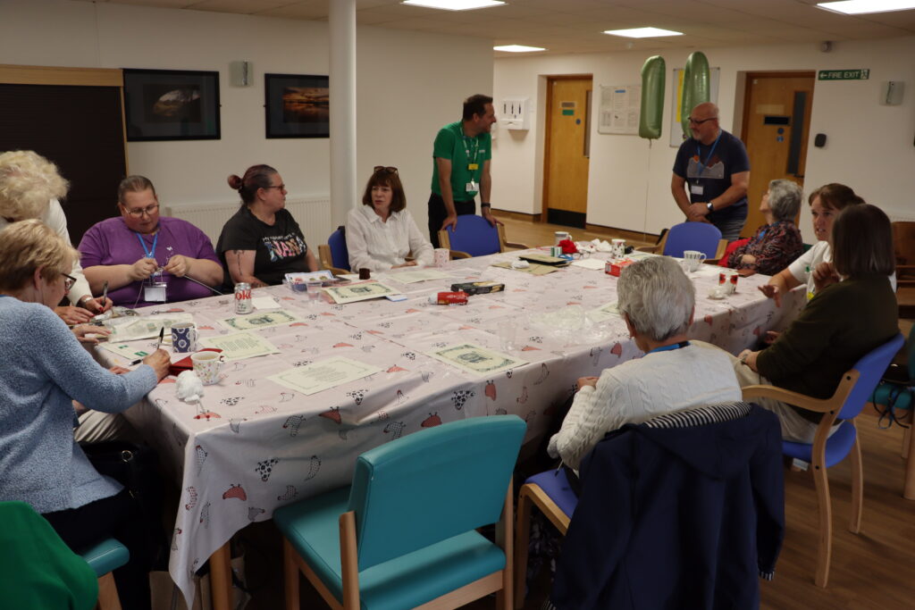The group sit around a table chatting, doing various crafts. These include knitting and embroidery.