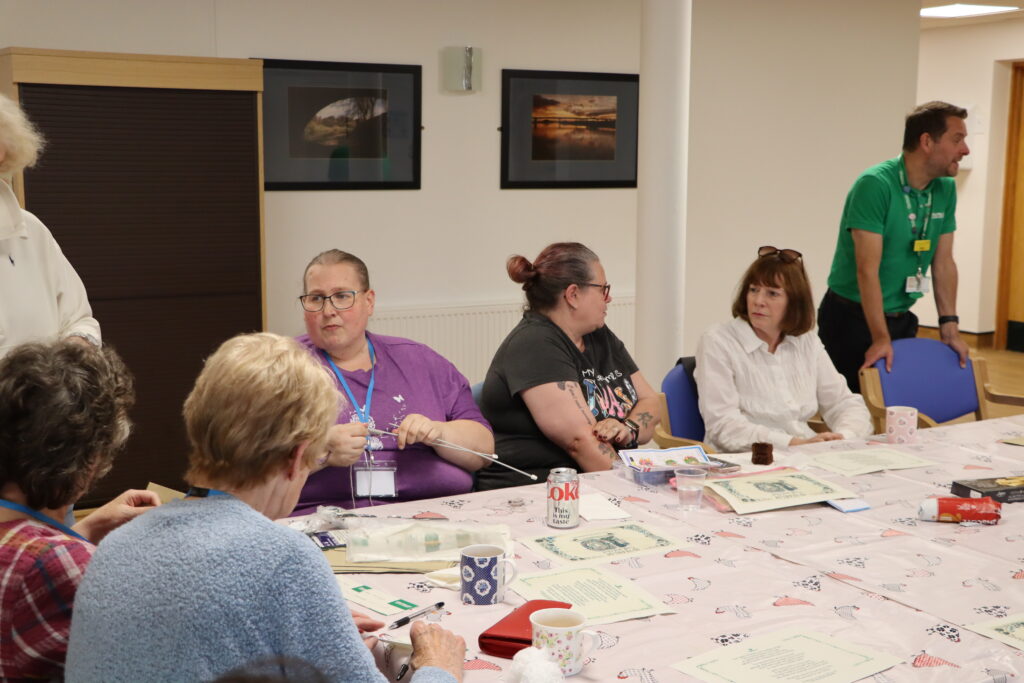 The group sit around a table chatting, doing various crafts. These include knitting and embroidery.
