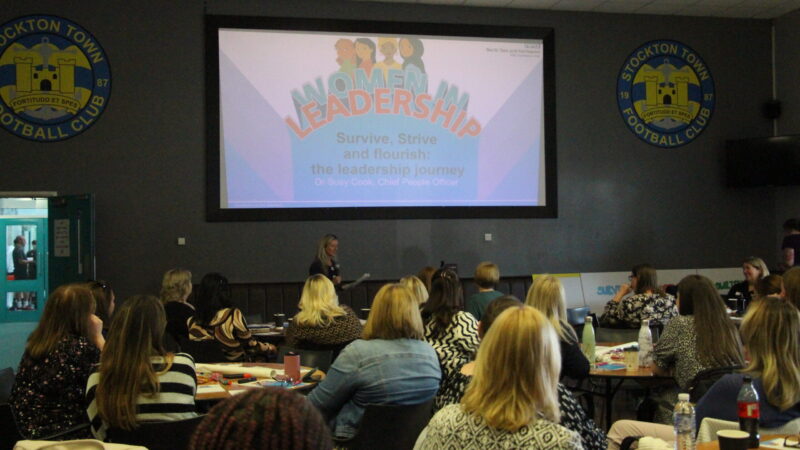 Susy Cook presents to an audience. A screen behind her reads: "Women in Leadership. Survive, strive and flourish: the leadership journey".