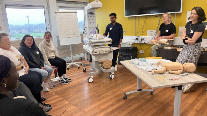 A group of people sit in discussion. Baby manikins lay on a table.