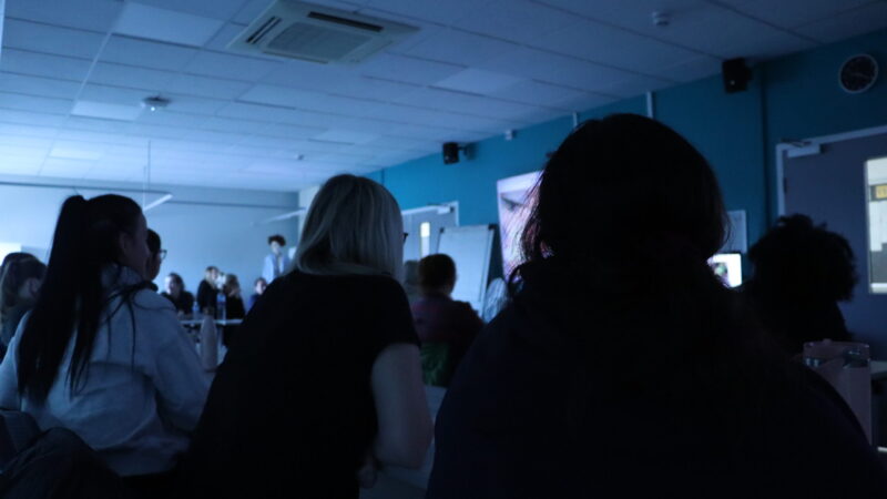 Students watch a large screen.