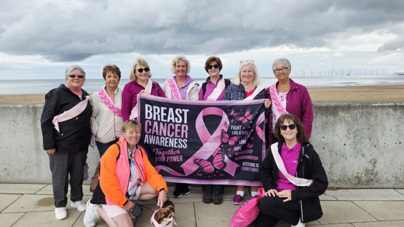 The walking group stand on the seafront. They hold a banner which reads: "Breast cancer awareness. Together, pink power".