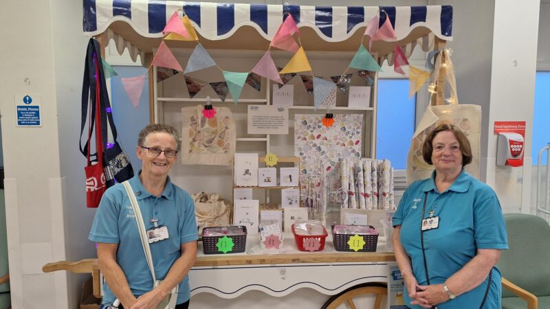 Two volunteer stand in front of a stall. The stall includes wrapping paper, greeting cards and tote bags.