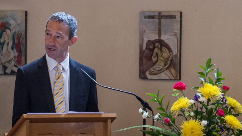 Neil Schneider stands at a podium. He is talking into a microphone.