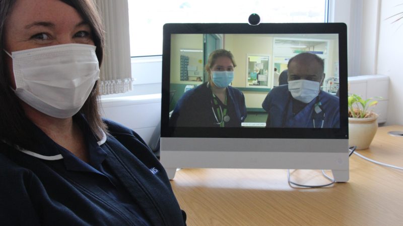 A nurse sits in front of an iPad. the iPad shows two doctors on screen.