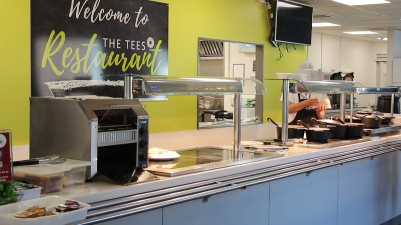 A food counter. Behind the counter a sign reads "Welcome to the Tees Restaurant".