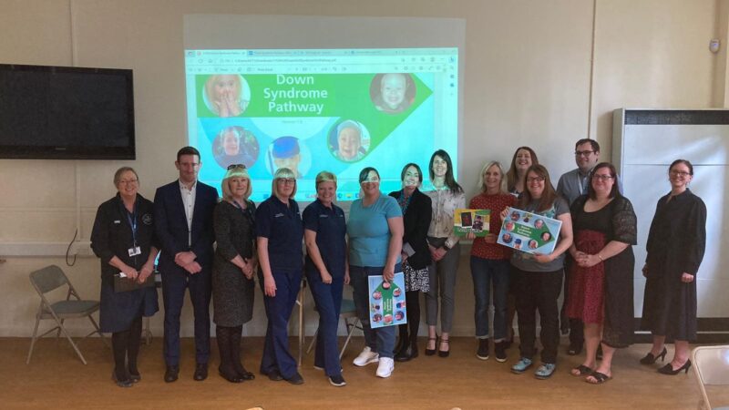 NHS staff and local parents stand in front of a screen. The screen reads: 'Down Syndrome Pathway' and shows images of children.