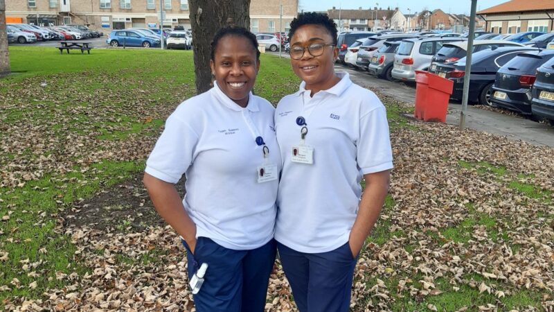 Rebecca Adione and Margaret Chigozie stand outside. They wear white polo shirts that read 'team support worker'.