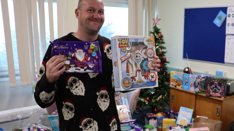 Volunteer Paul Willows stands in front of a pile of food and toy donations. We holds a selection box and Toy Story toy.