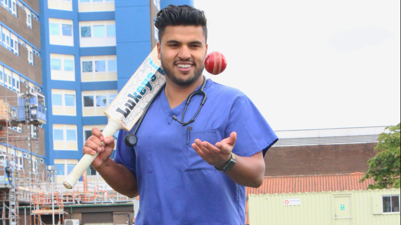 Vishal stands outside the University Hospital of North Tees. He is holding a cricket bat and throwing a ball in the air.