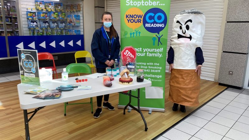 Leanne Watson at a stand. She has information leaflets and props about smoking. Someone stands next to her wearing a costume of a cigarette with an angry face.