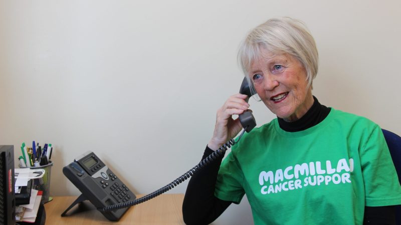 A volunteer sitting at a desk. She is speaking on the phone. Her t-shirt reads: "Macmillan cancer support".