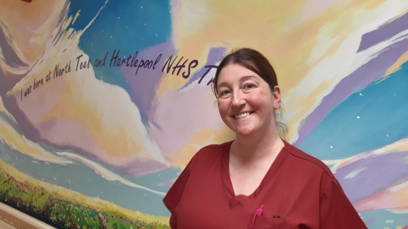 Nicola Riley stands in the corridor in front of a wall mural. of a field. The mural reads: "I wan born at North Tees and Hartlepool NHS Trust".