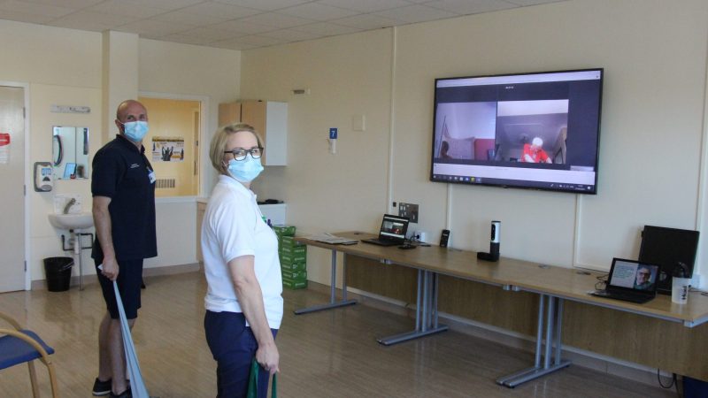 Two members of the team on a virtual call with two patient. The patients are shown on a big screen on the wall.