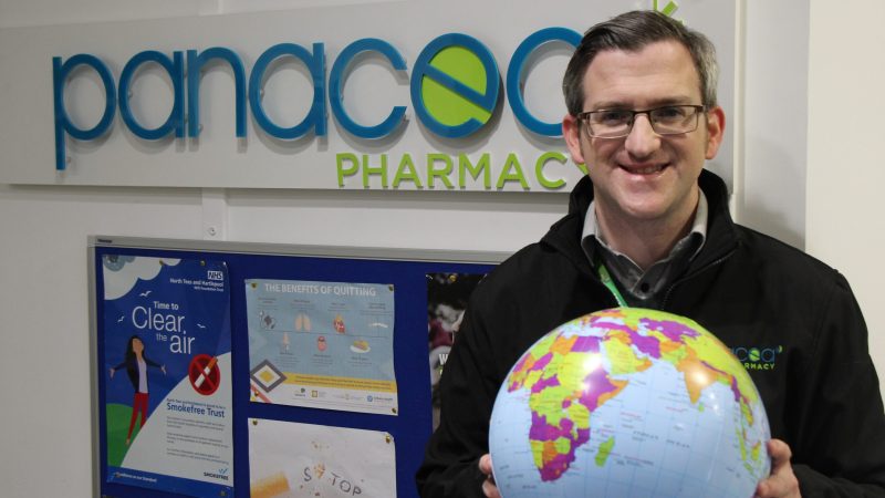 Patrick Russell stands in front of a Panacea sign. He holds a blow up globe.