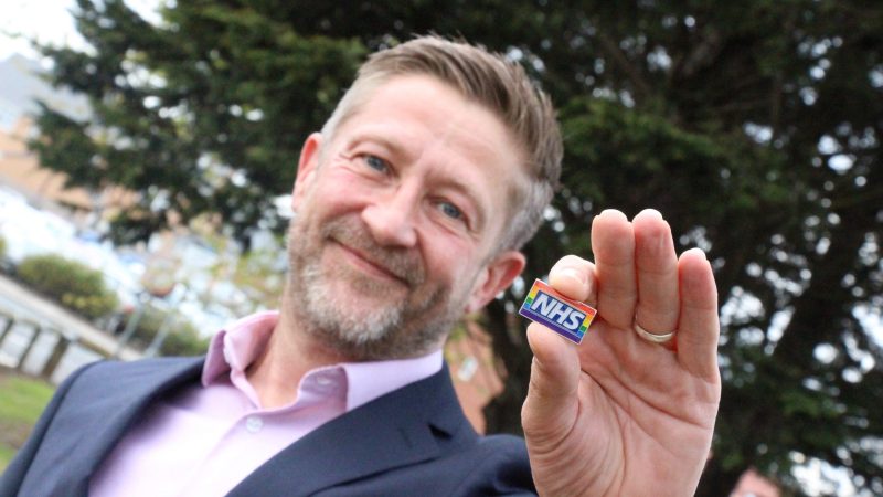 Levi Buckley holds an NHS rainbow badge. It is a Pride flag with the NHS logo on it.