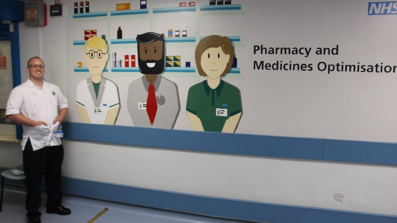Mark Walmsley stands in front of his painting. The painting shows three cartoon pharmacists in front of medication shelves.